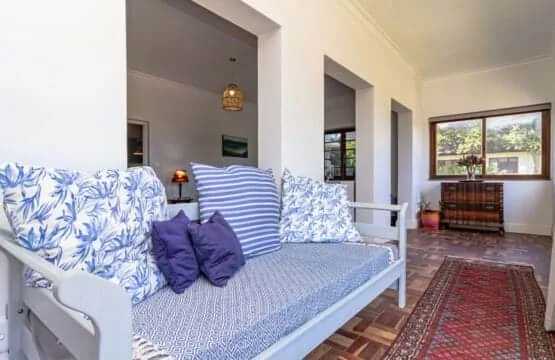 Una cama de día blanca con cojines azules estampados está junto a una alfombra roja en una habitación luminosa.