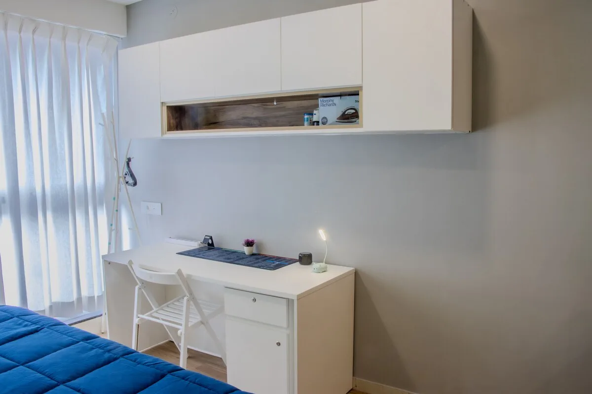 Un espacio de trabajo en un dormitorio, con escritorio y armarios blancos junto a una cama azul.