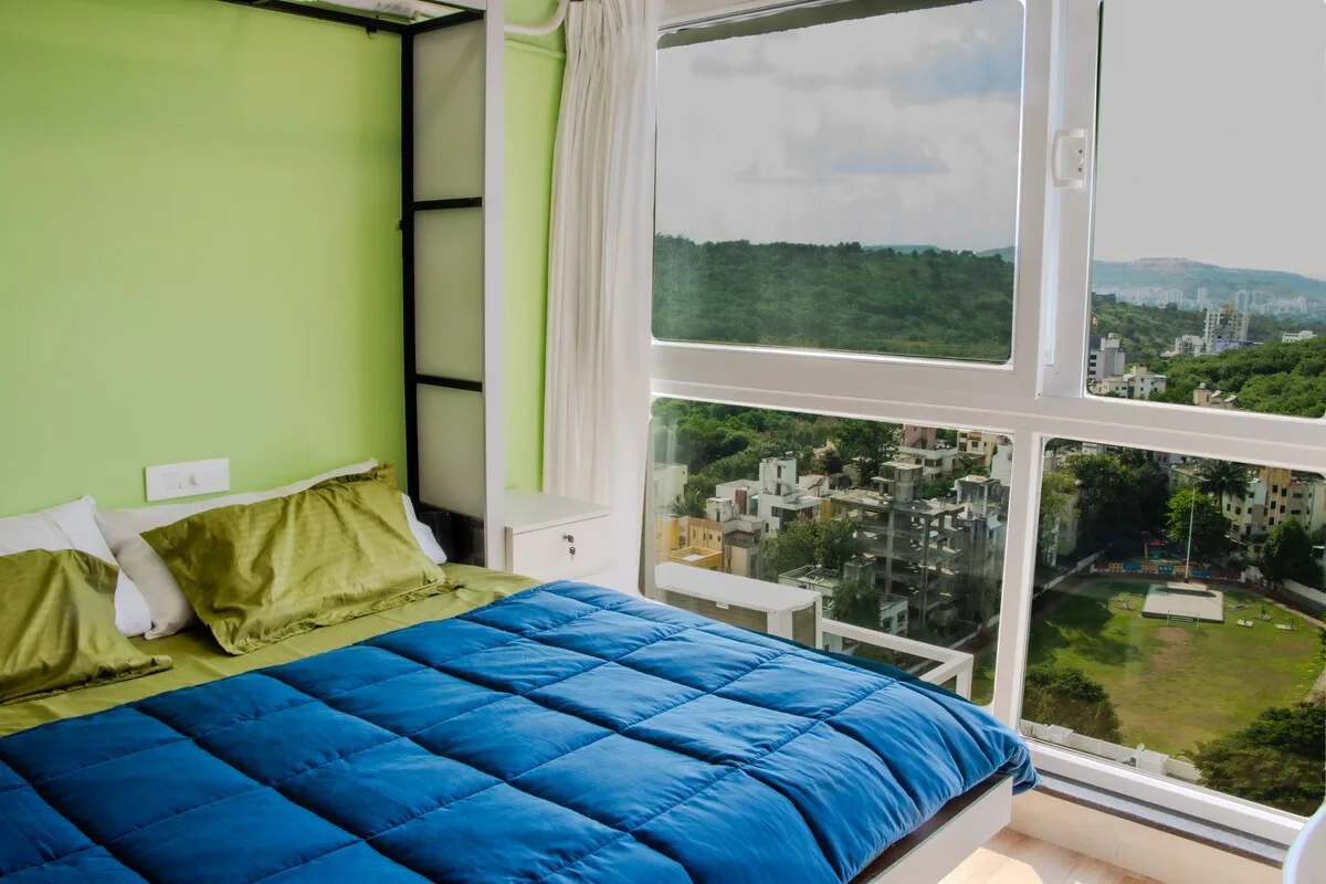 Un dormitorio con una cama junto a grandes ventanas con vistas a vegetación y edificios.