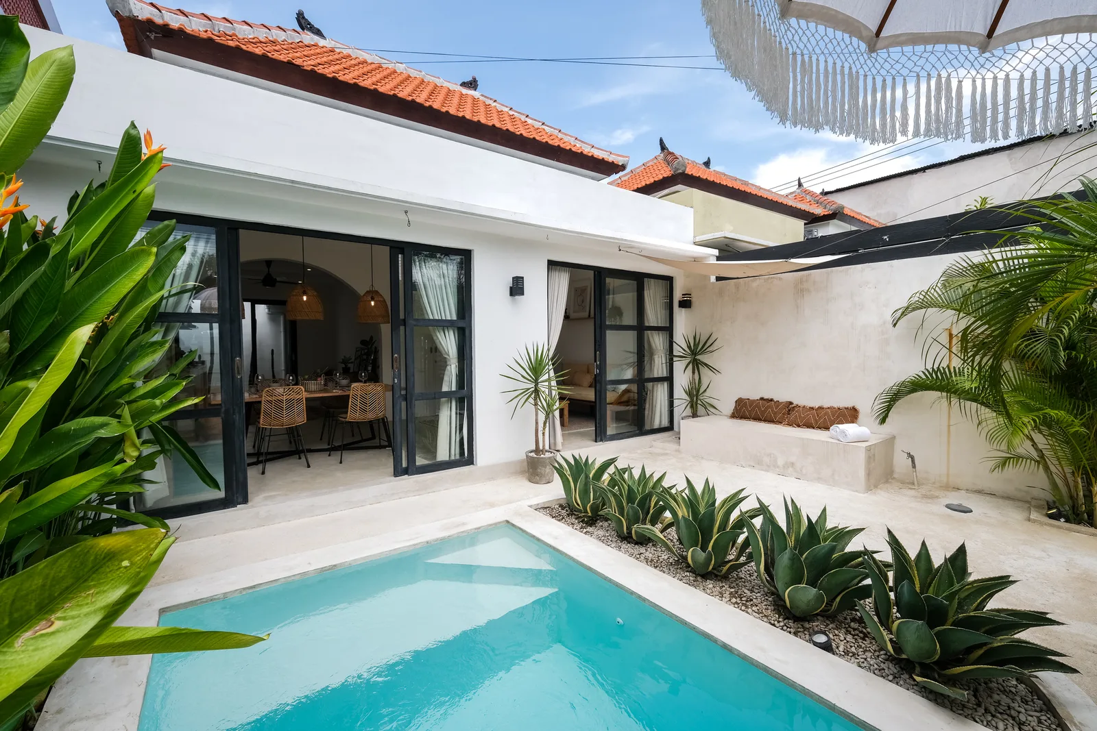 Una piscina en un patio junto a un edificio blanco, con plantas tropicales y un comedor al aire libre.