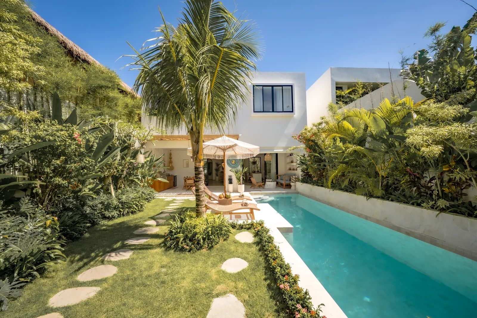 Una villa blanca moderna da a una piscina turquesa, rodeada de una palmera, vegetación y tumbonas.