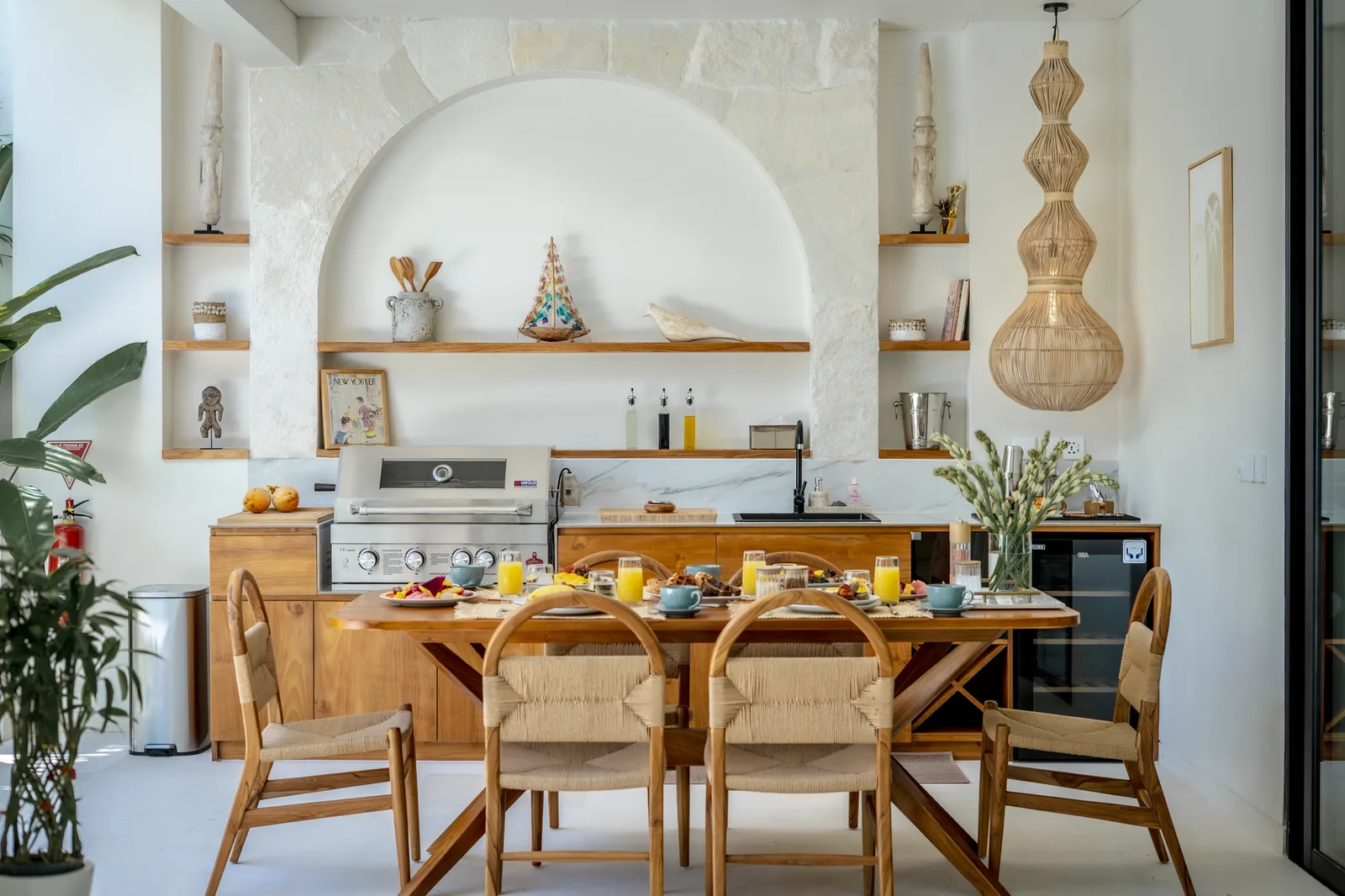 Un comedor luminoso cuenta con una mesa de madera, sillas tejidas y una cocina con estantes abiertos en arco.