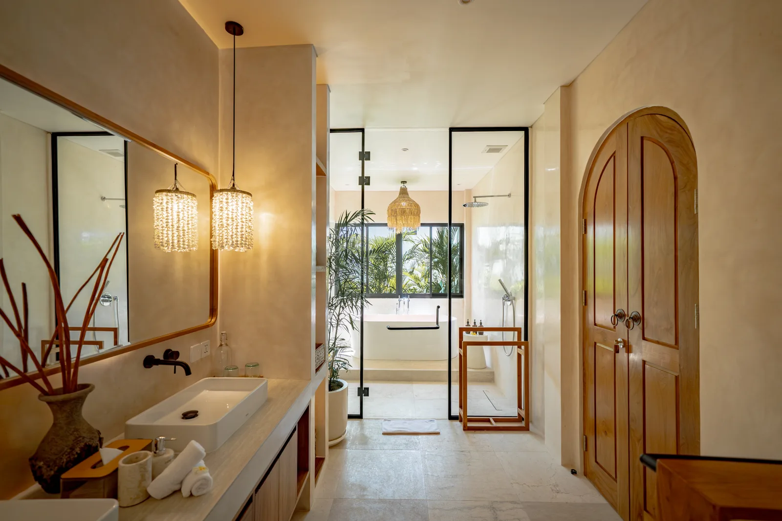 Un lavabo de baño con lavamanos sobre encimera, dos lámparas colgantes y una ducha acristalada junto a una puerta de madera arqueada.