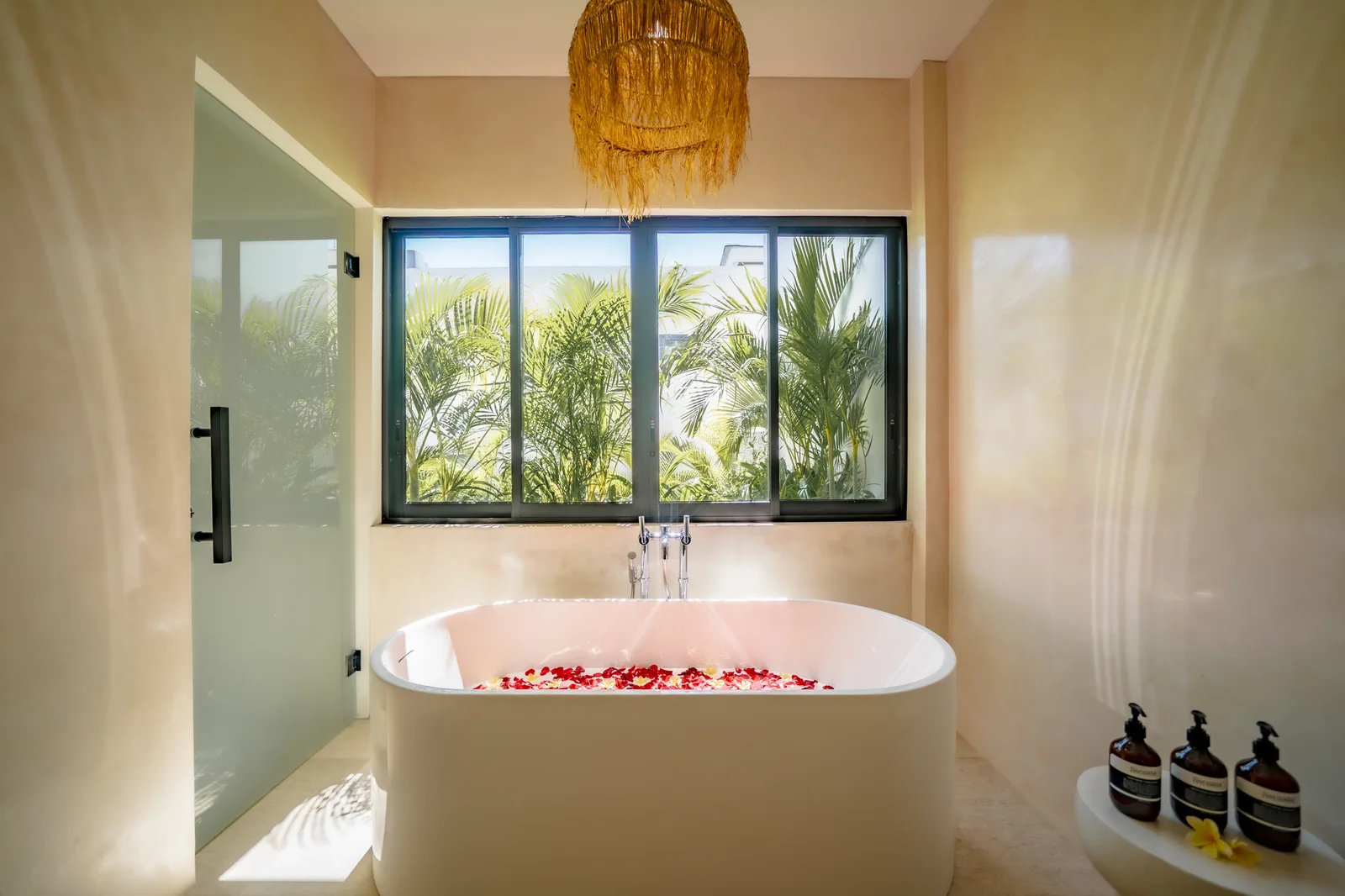 Una bañera exenta está bajo una amplia ventana con palmeras al fondo y una lámpara colgante de fibras.