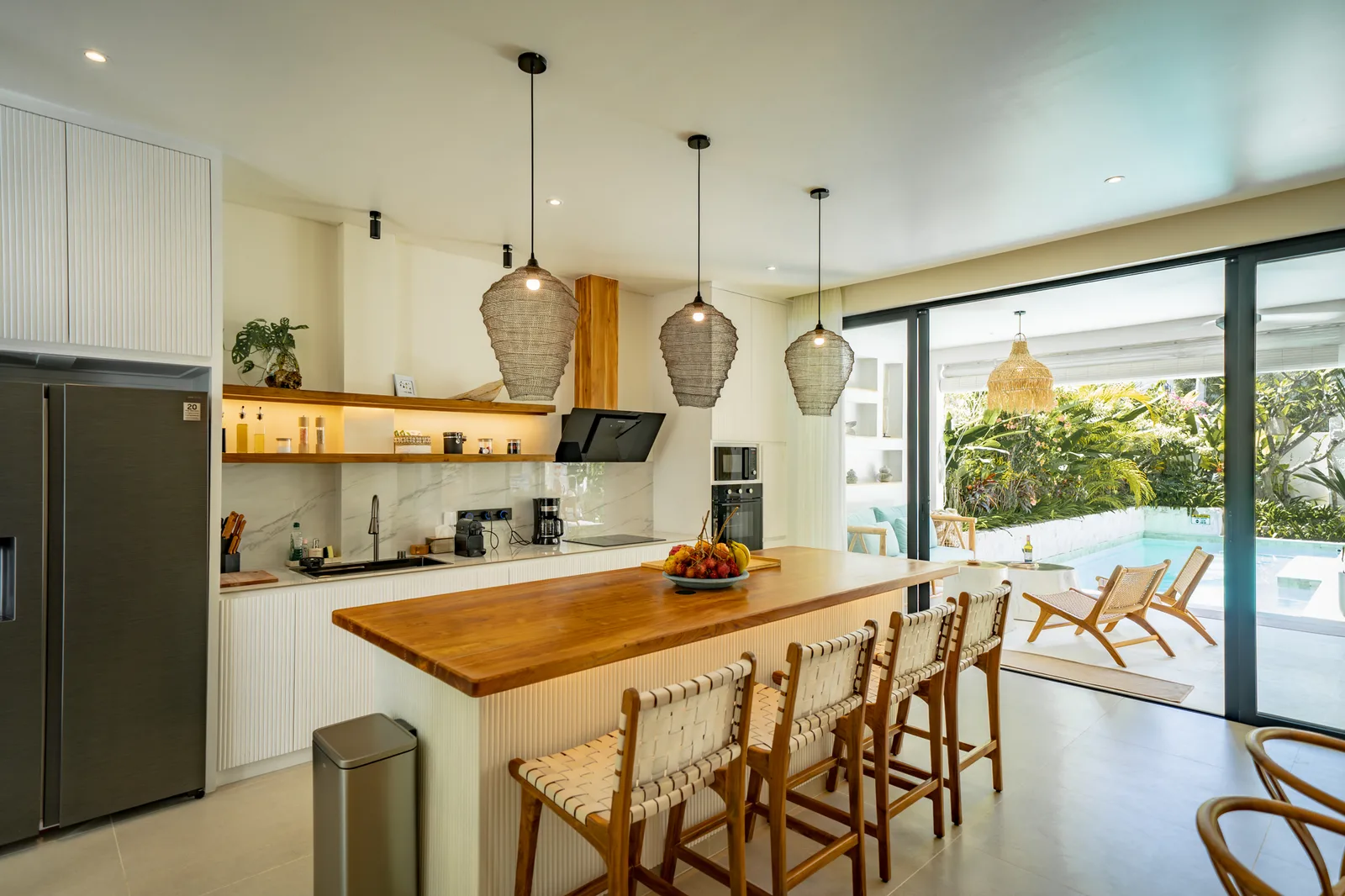Una cocina abierta y luminosa cuenta con una larga isla de madera, taburetes tejidos, lámparas colgantes y puertas de vidrio al patio.