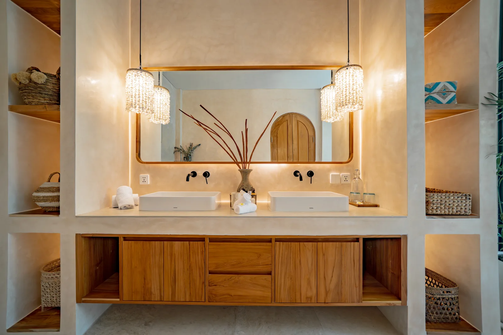 Un tocador de baño con dos lavabos está bajo un espejo amplio, con gabinetes de madera y estantes laterales empotrados.