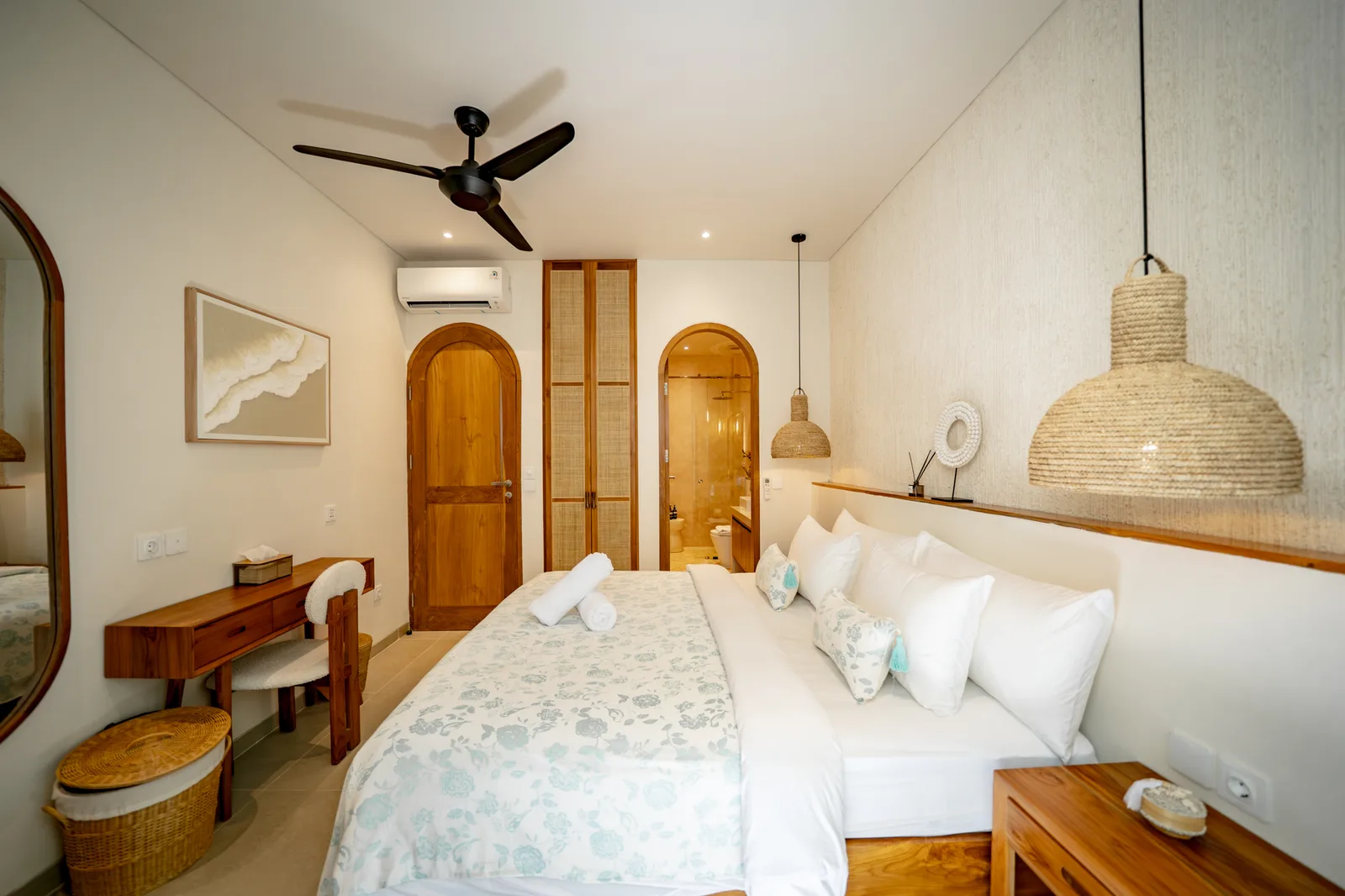 Un dormitorio luminoso con cama blanca, ropa de cama estampada clara, puertas de madera y lámparas colgantes trenzadas.