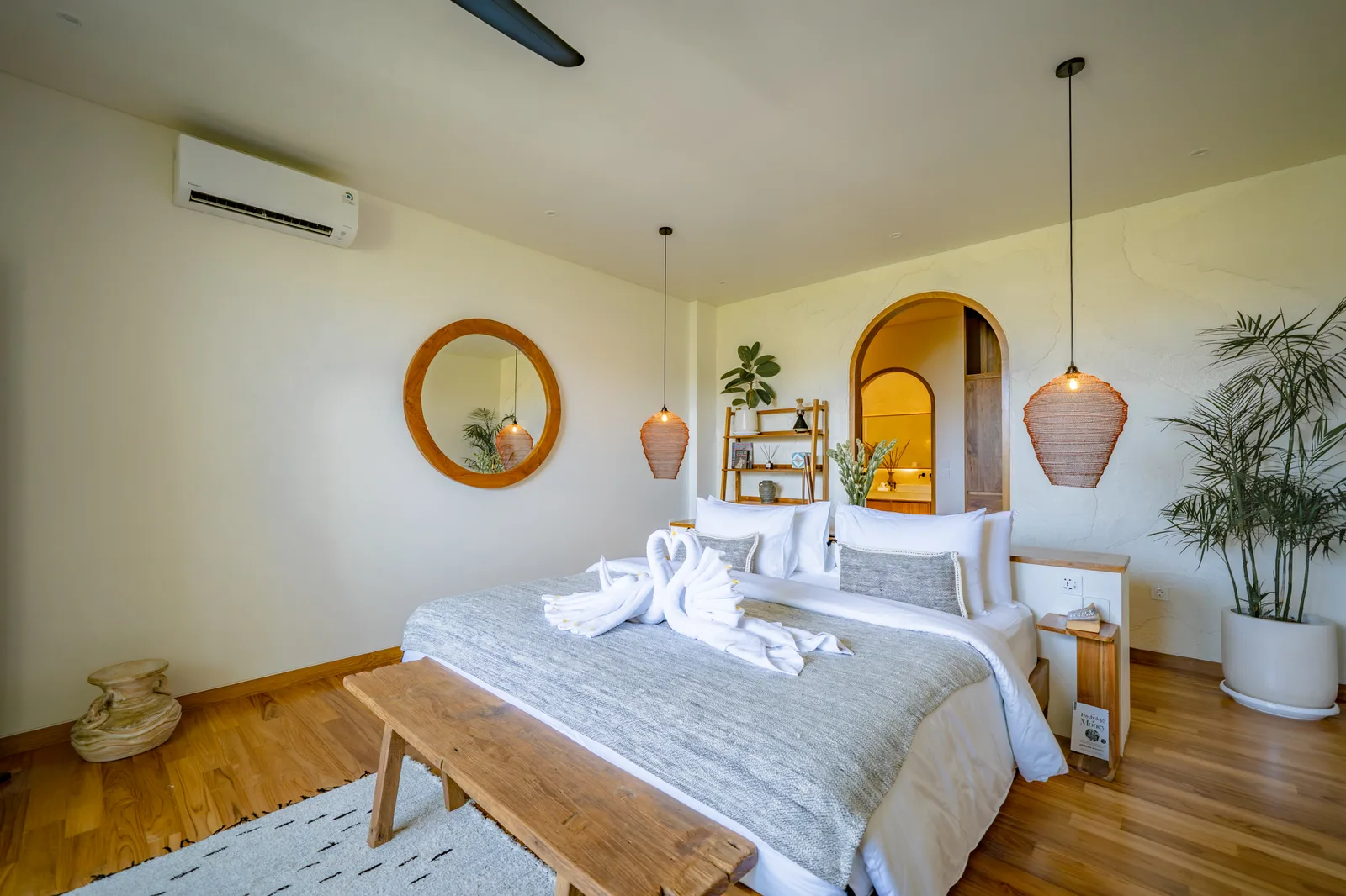 Un dormitorio luminoso con una cama bien tendida, un espejo redondo, lámparas colgantes y plantas.