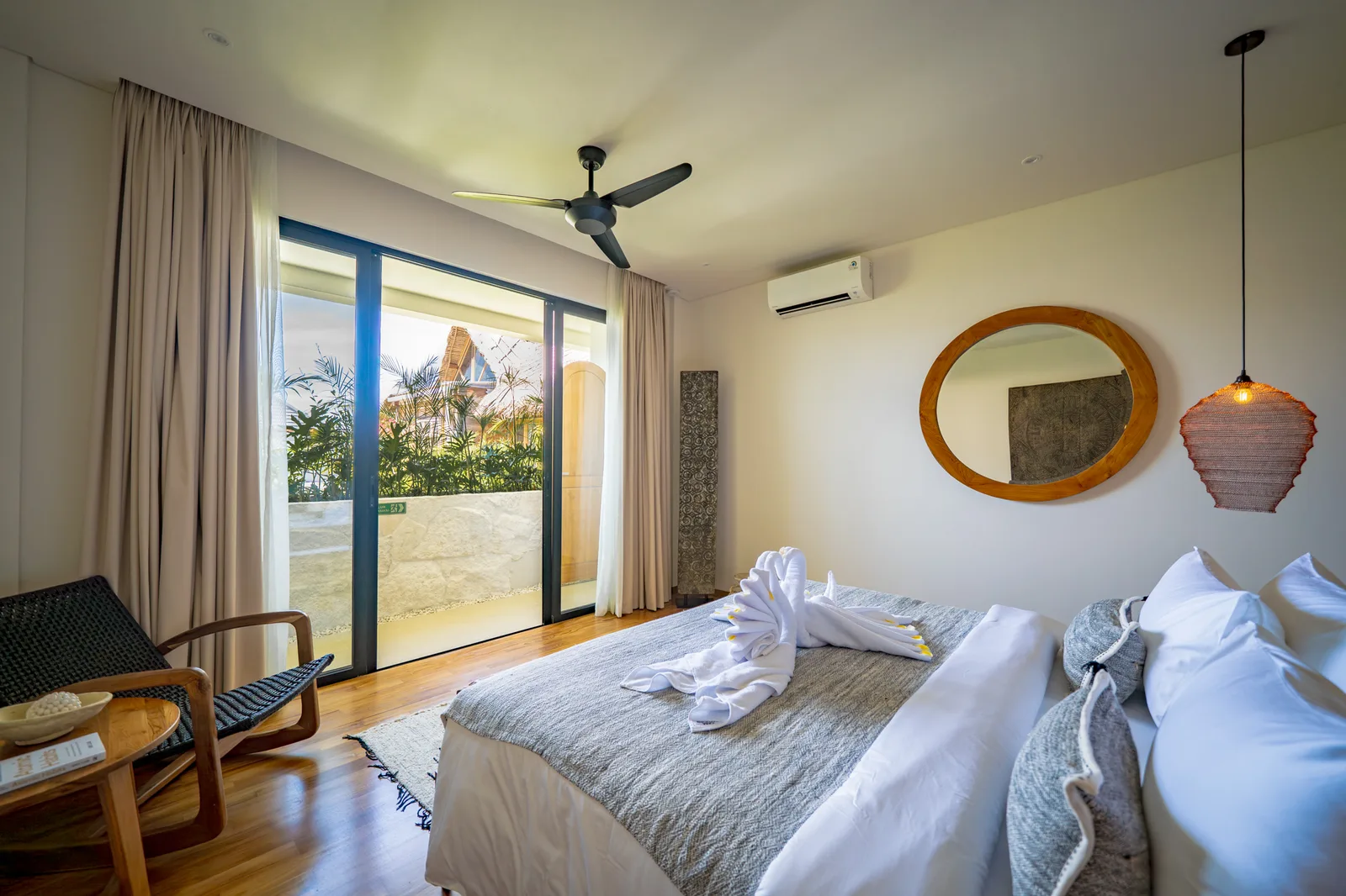 Un dormitorio con una cama blanca, toallas dobladas, sillas de madera y puertas corredizas de vidrio hacia un patio.