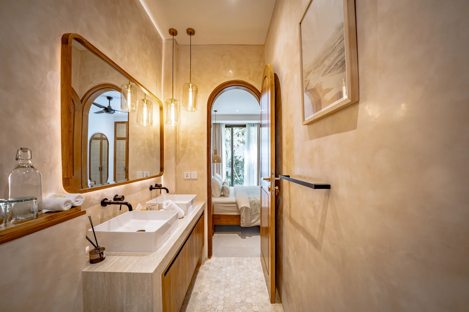 Un baño luminoso tiene un tocador largo con lavabo sobre encimera, un espejo grande y una puerta arqueada hacia un dormitorio.