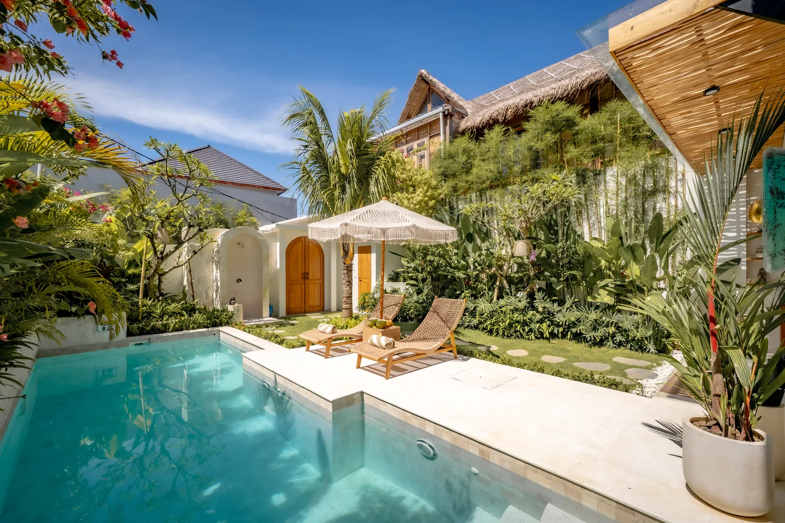 Un patio tropical cuenta con una piscina turquesa, dos tumbonas, una sombrilla y abundante vegetación.
