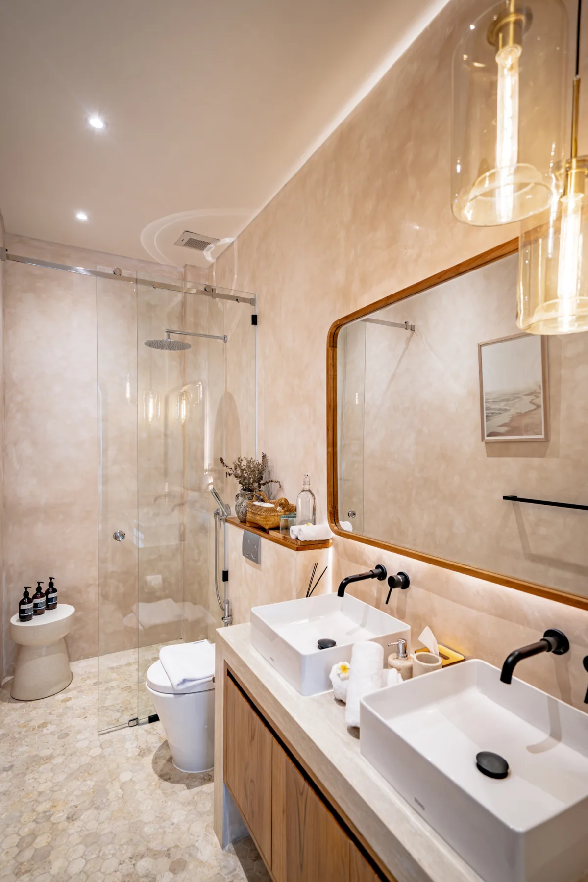Un baño moderno cuenta con una ducha de vidrio, dos lavabos sobre encimera y un largo espejo con marco de madera.