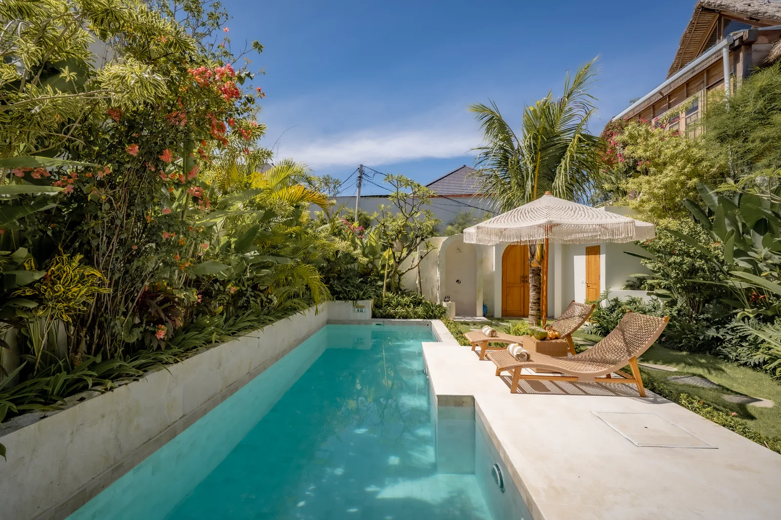 Una piscina estrecha de color turquesa bordea un patio blanco con tumbonas, una sombrilla y exuberantes plantas tropicales.