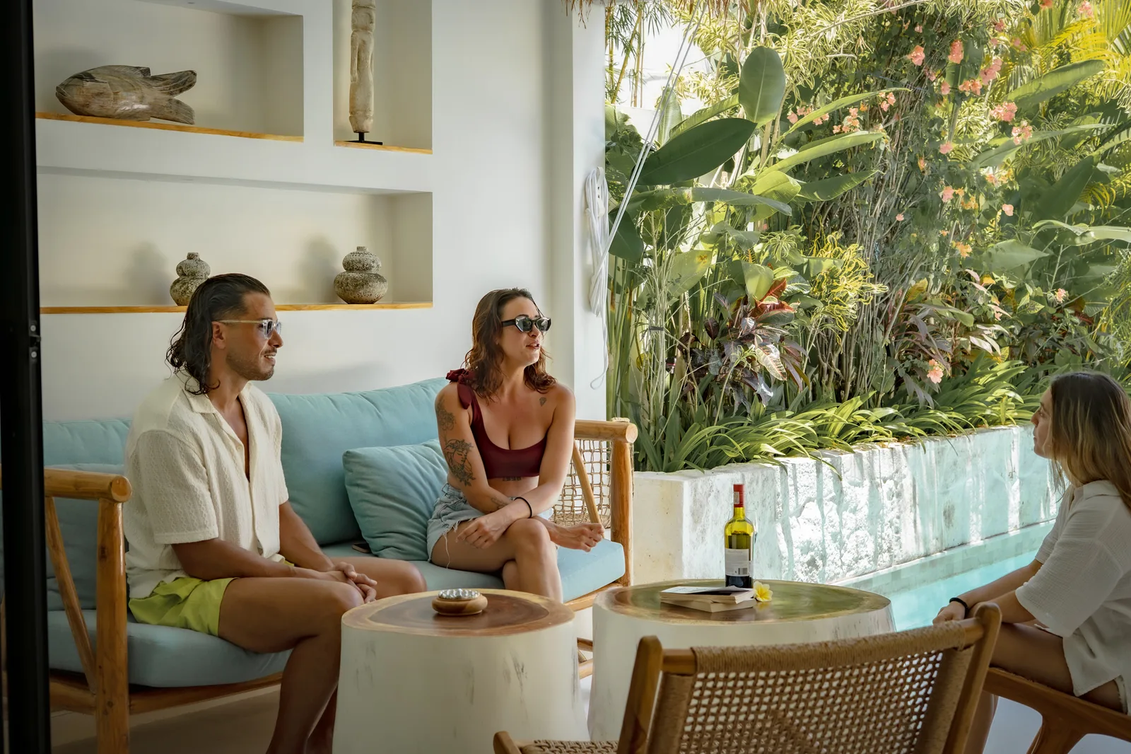 Tres personas se sientan alrededor de una mesa baja junto a un gran ventanal con vistas a plantas tropicales.