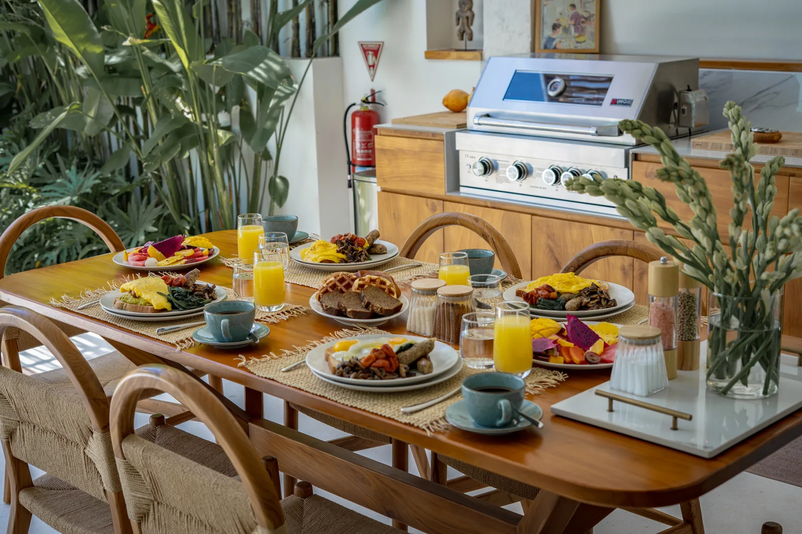 Una mesa de comedor está servida con platos de desayuno, café, zumo y flores, junto a una cocina.