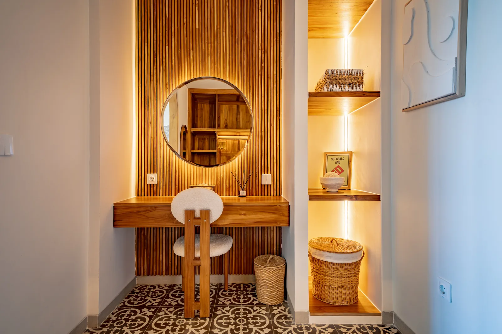 Un tocador de madera con espejo redondo y silla está junto a estantes iluminados y una cesta tejida.