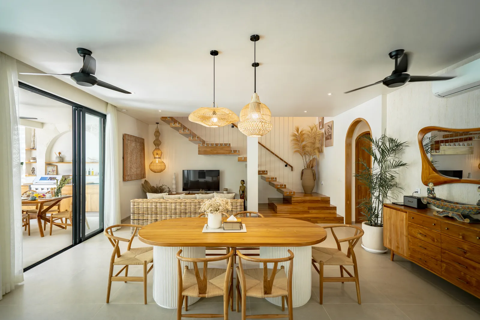 Una sala abierta con comedor y estar presenta una mesa de madera, lámparas colgantes tejidas y escaleras a un nivel superior.