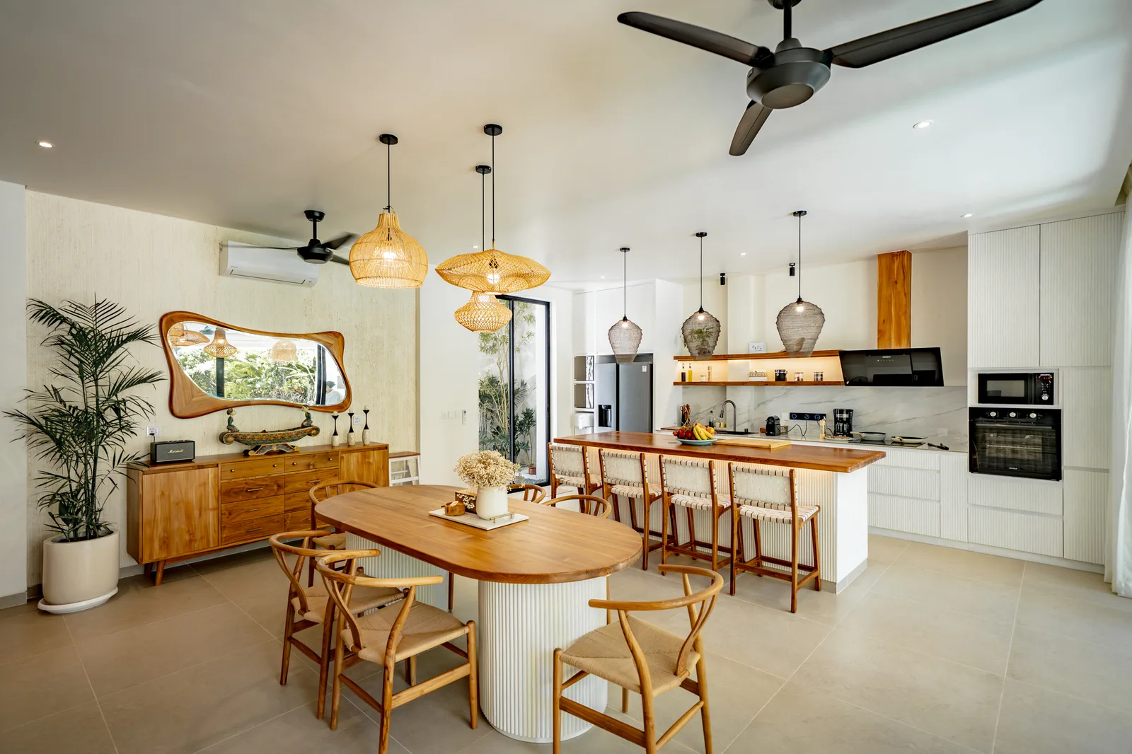 Una luminosa cocina-comedor diáfana cuenta con una larga mesa de madera, lámparas colgantes tejidas y armarios blancos.