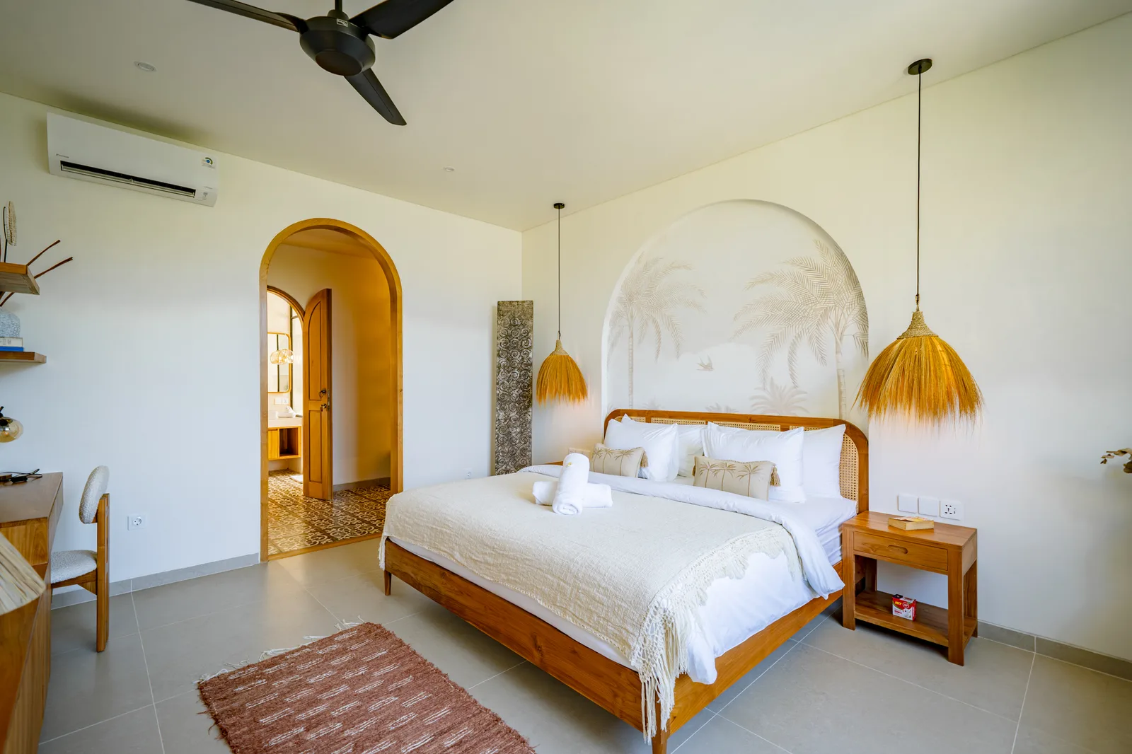 Un dormitorio luminoso cuenta con una cama de madera, ropa de cama blanca, lámparas colgantes tejidas y una puerta arqueada.