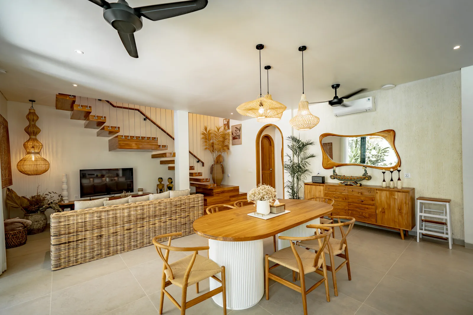 Una luminosa sala de estar y comedor tiene una mesa de madera, sillas tejidas, lámparas colgantes y una escalera.