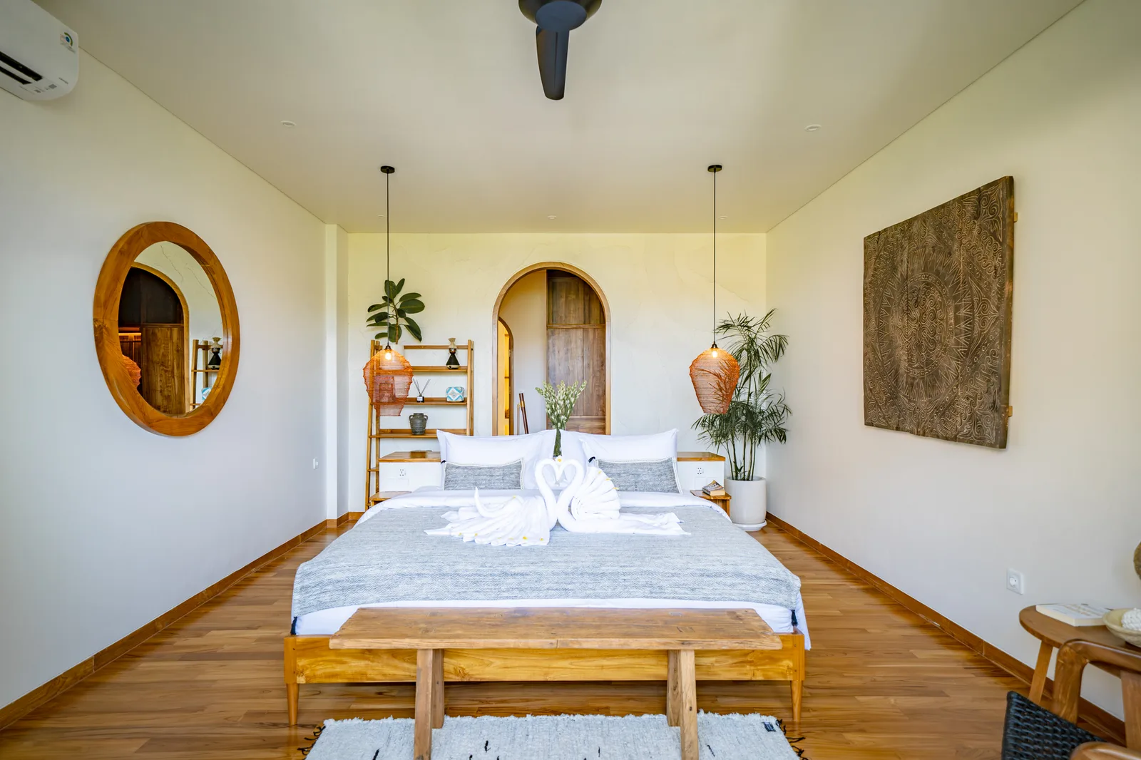 Un dormitorio luminoso con una cama grande, un banco de madera, un espejo arqueado y plantas en macetas.