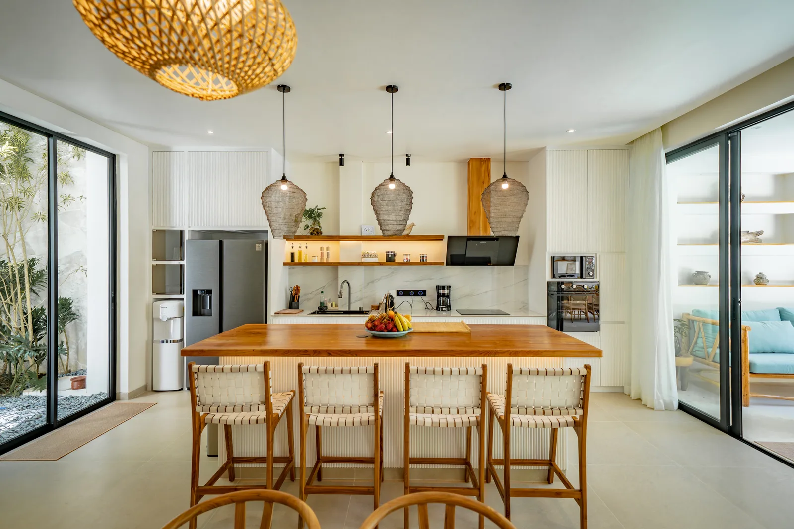 Cocina luminosa de planta abierta con una larga isla de madera, lámparas colgantes trenzadas y puertas de vidrio al patio.
