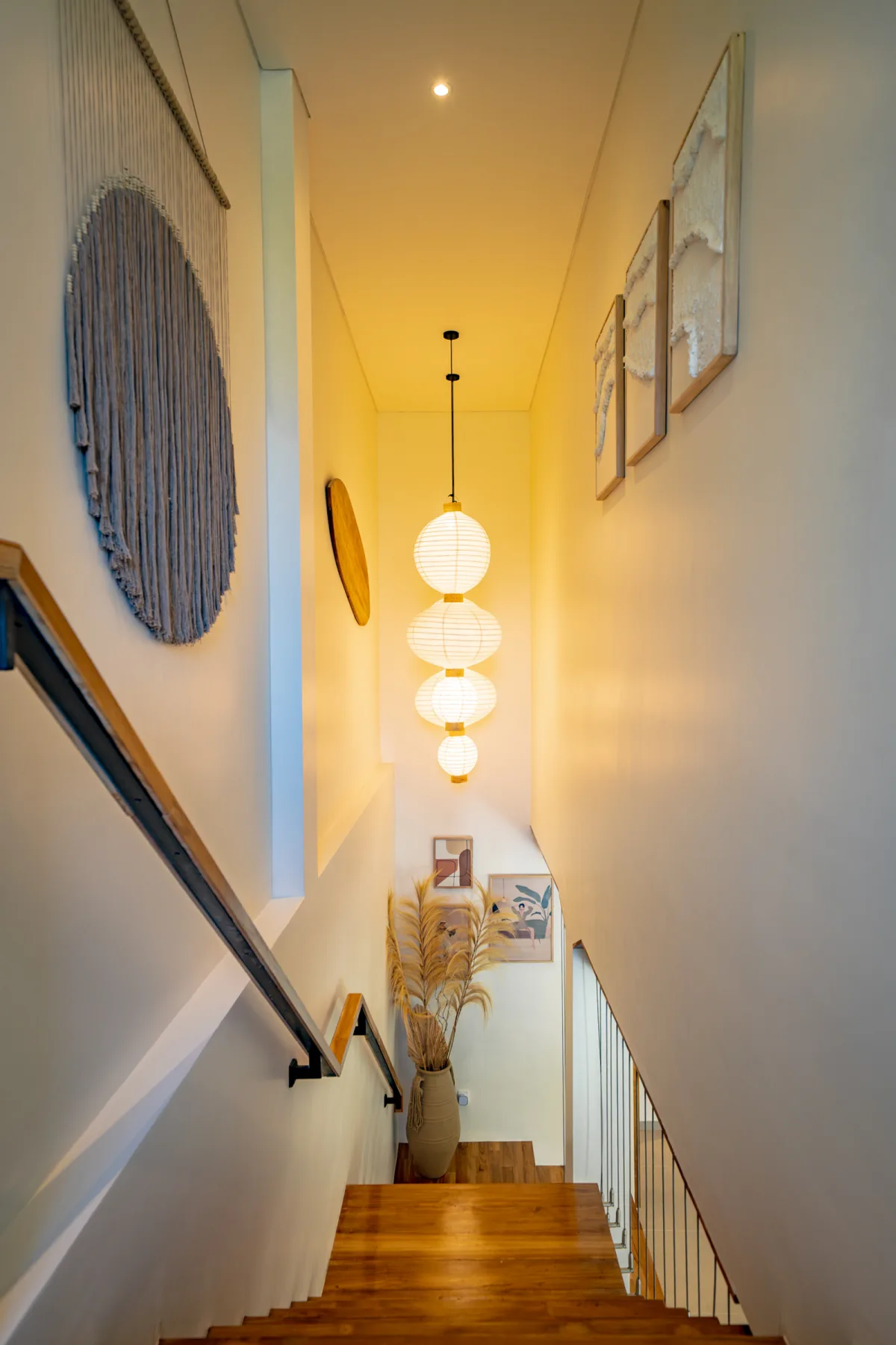 Una escalera estrecha con peldaños de madera, pasamanos negro, cuadros y lámparas colgantes encendidas.