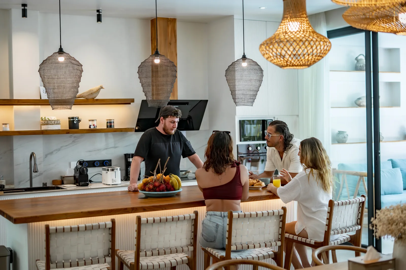 Una cocina luminosa con una isla y lámparas colgantes de mimbre, donde tres personas se sientan mientras un hombre prepara comida.