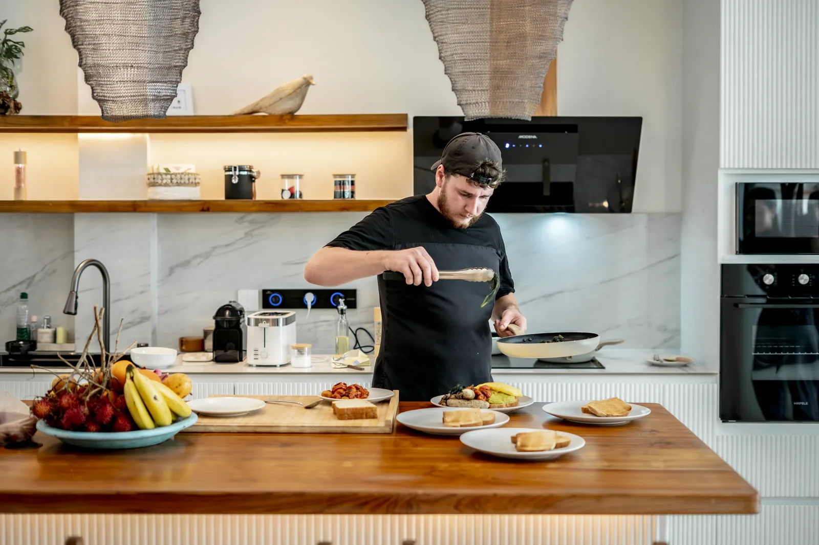 Un hombre prepara comida en una isla de cocina, con fruta, platos servidos y electrodomésticos integrados cerca.