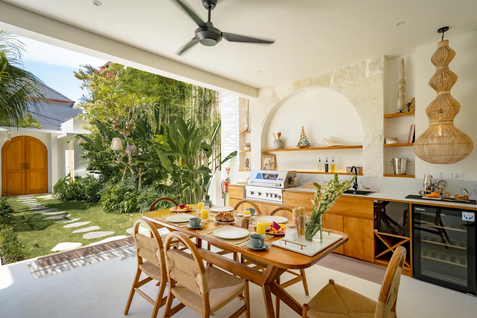 Una luminosa cocina y comedor al aire libre cuentan con armarios de madera, una mesa larga y abundante vegetación exterior.
