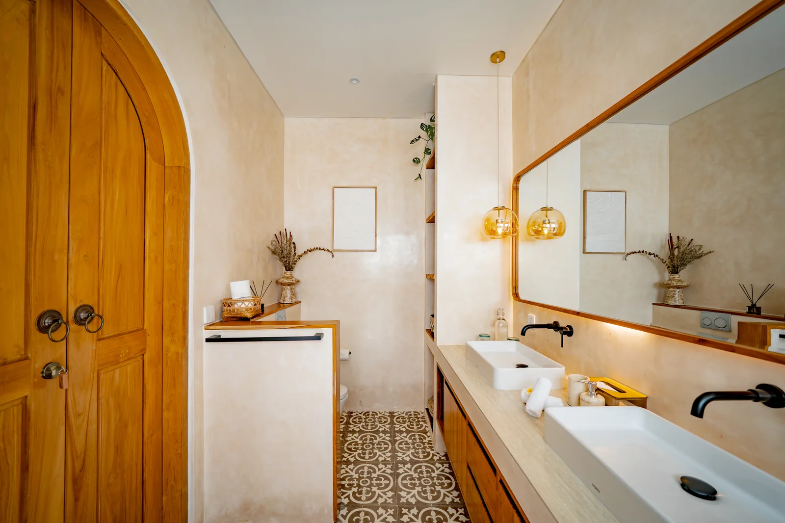 Un baño luminoso tiene un largo tocador doble, un espejo grande, suelo de baldosas estampadas y una puerta de madera arqueada.