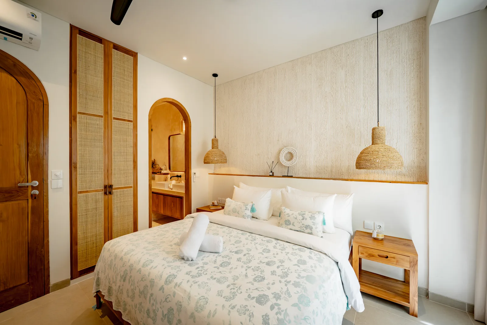 Un dormitorio luminoso con una cama grande, lámparas colgantes de fibra tejida y puertas y muebles de madera cálida.