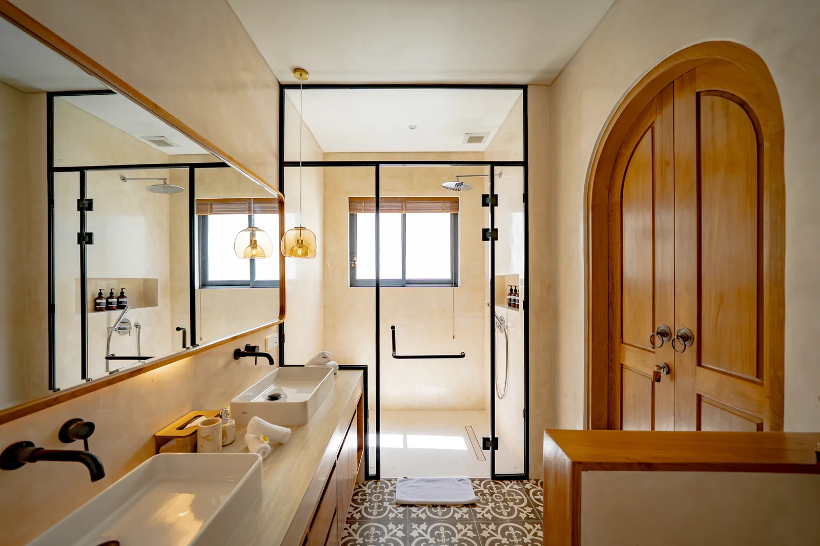 Un baño luminoso con ducha de vidrio, dos lavabos y una puerta de madera redondeada.