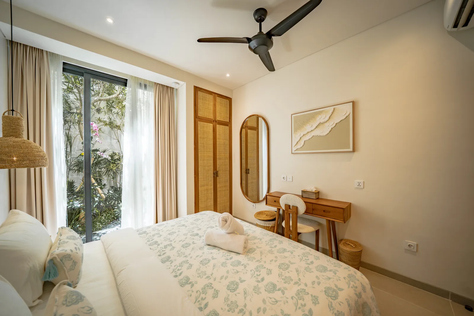 Un dormitorio luminoso tiene una colcha floral, puertas de vidrio hacia la vegetación y un pequeño escritorio bajo un cuadro.