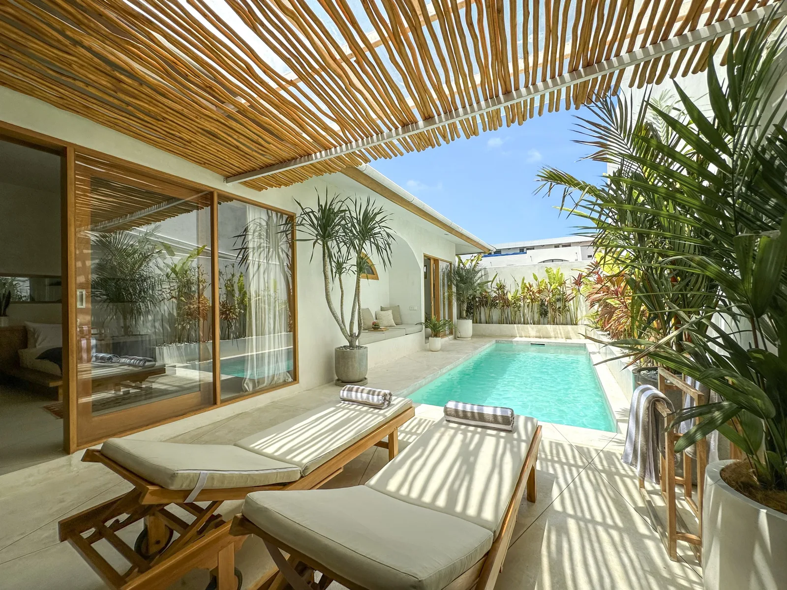 A sunlit pool patio with lounge seating beneath a slatted pergola.