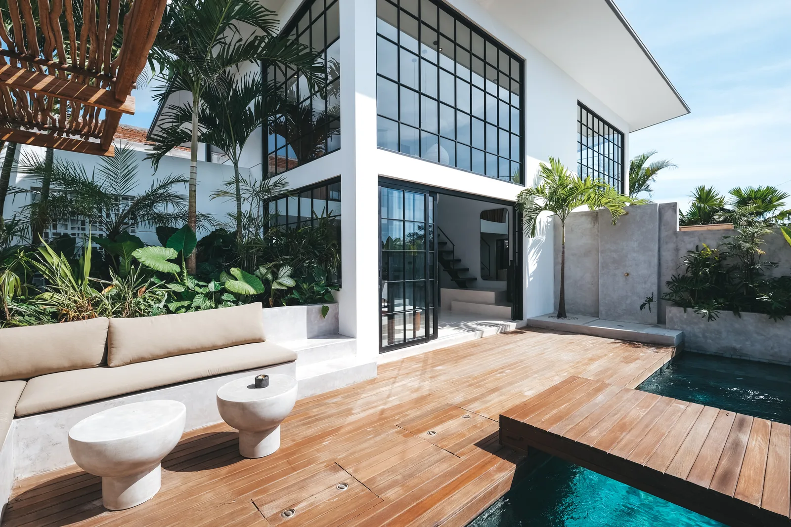 A modern house opens onto a wooden deck beside a swimming pool and outdoor seating.