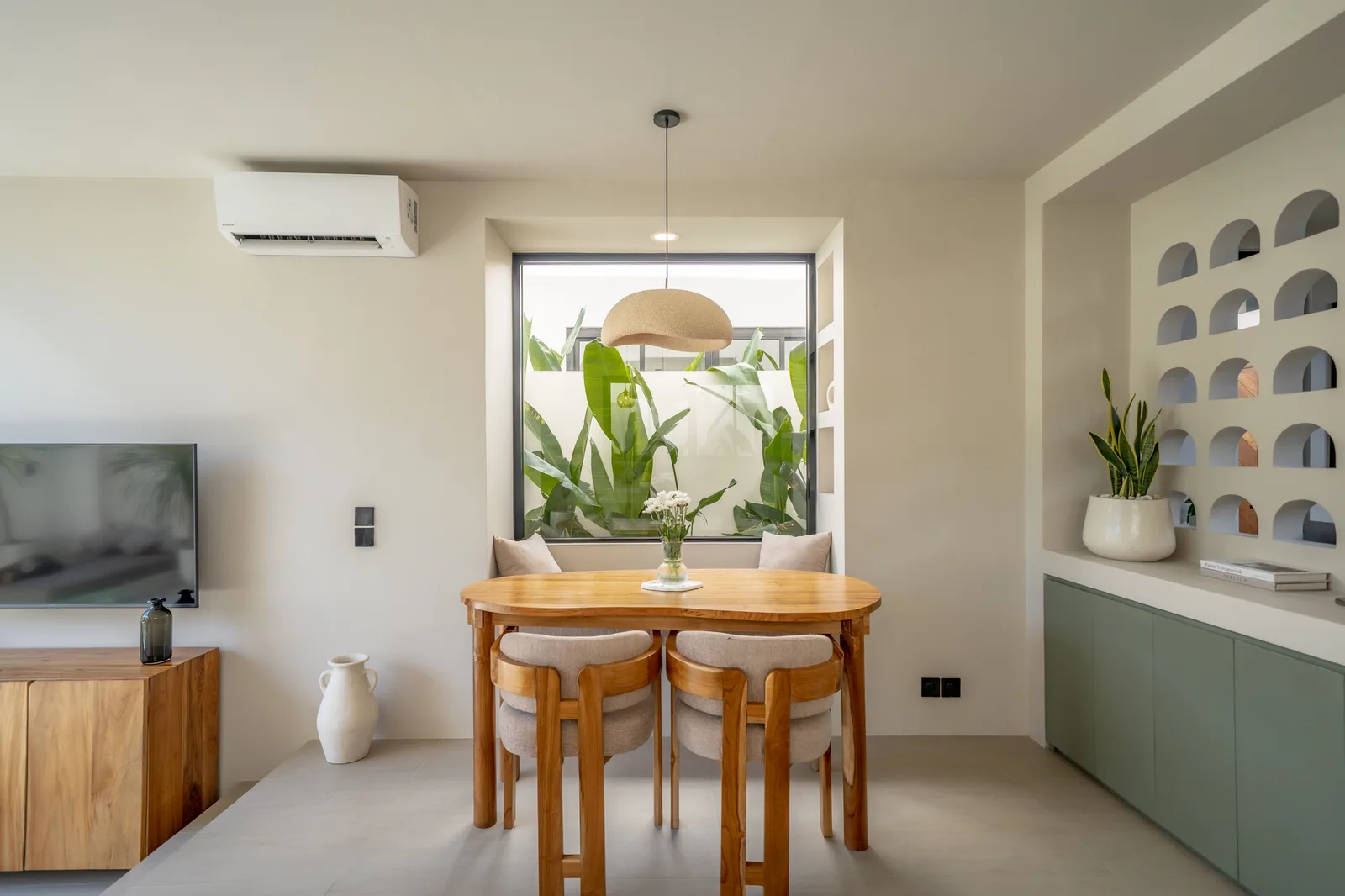 Una mesa de comedor con sillas está frente a una gran ventana llena de plantas, con un televisor y estantes empotrados cerca.