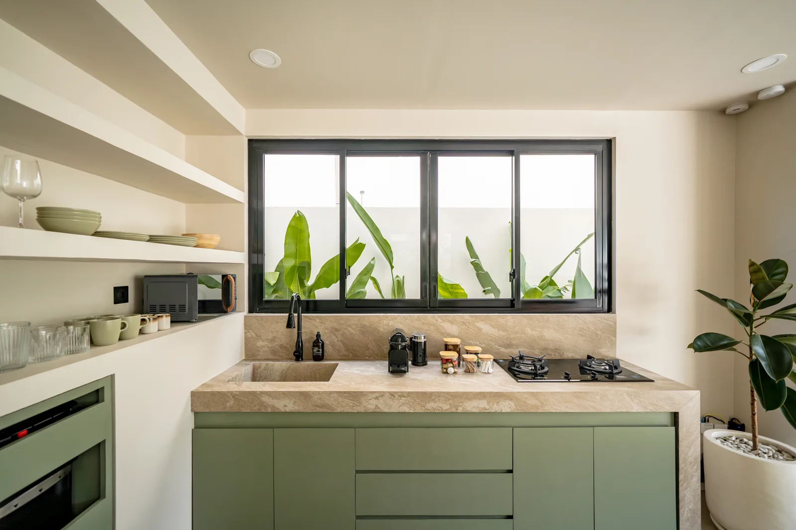 Una cocina moderna con gabinetes verde salvia, una encimera de piedra, una placa de cocina y ventanas amplias.