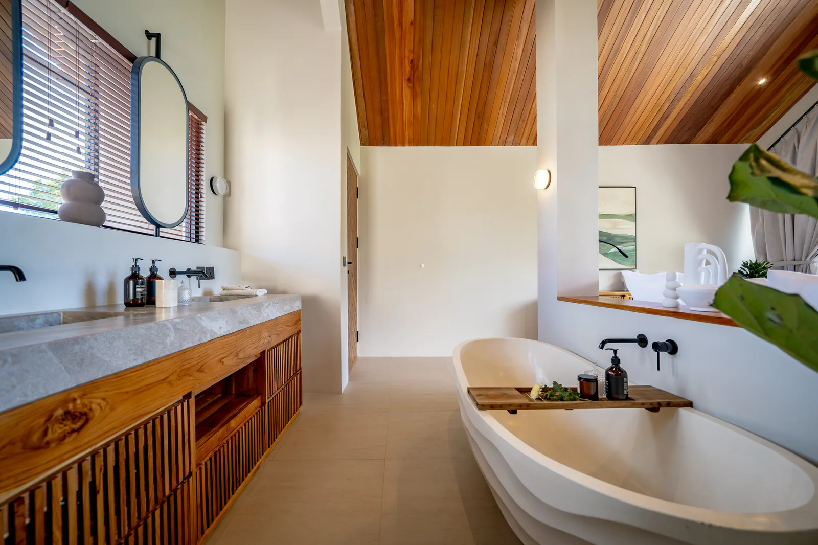 Un baño luminoso cuenta con un largo tocador de madera, espejo ovalado, bañera exenta y techo de madera cálida.