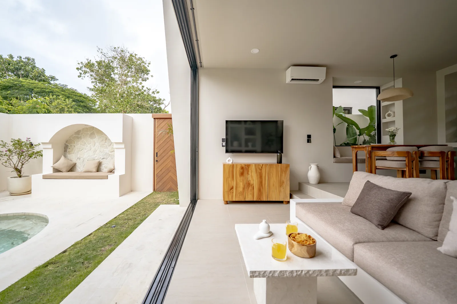 Una luminosa sala de estar de planta abierta con sofá gris, televisor, mueble de madera y comedor junto al jardín.