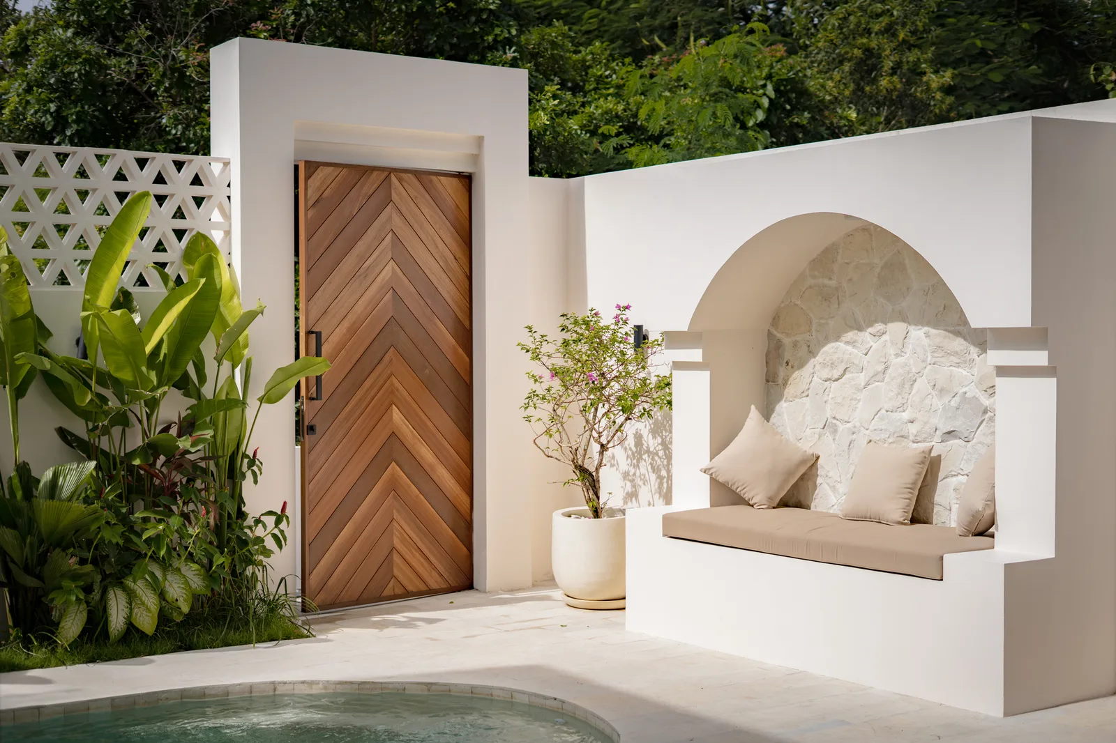 Un rincón de patio blanco tiene un banco empotrado, un arco con fondo de piedra y una puerta de madera junto a plantas frondosas.