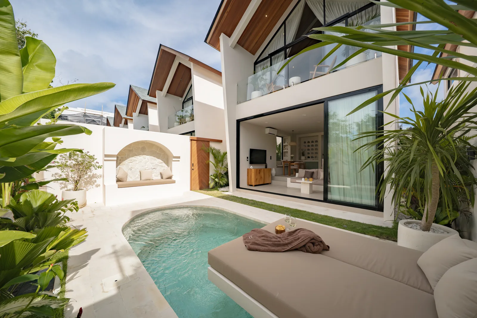 Una villa blanca moderna da a una pequeña piscina, con puertas de vidrio y plantas tropicales alrededor del patio.