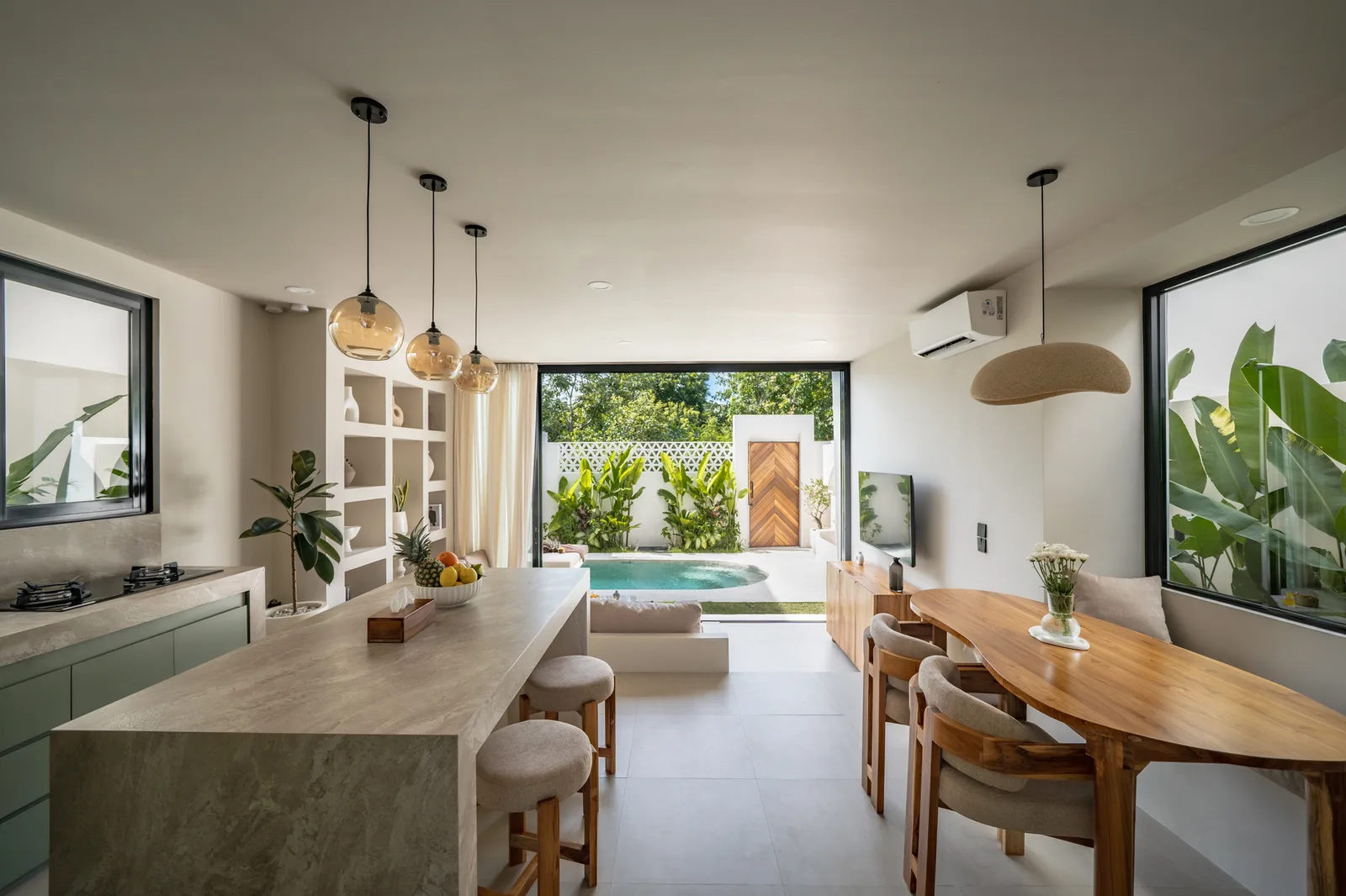 Una cocina y comedor de planta abierta tienen una larga isla de piedra, una mesa de madera y puertas amplias hacia el jardín.