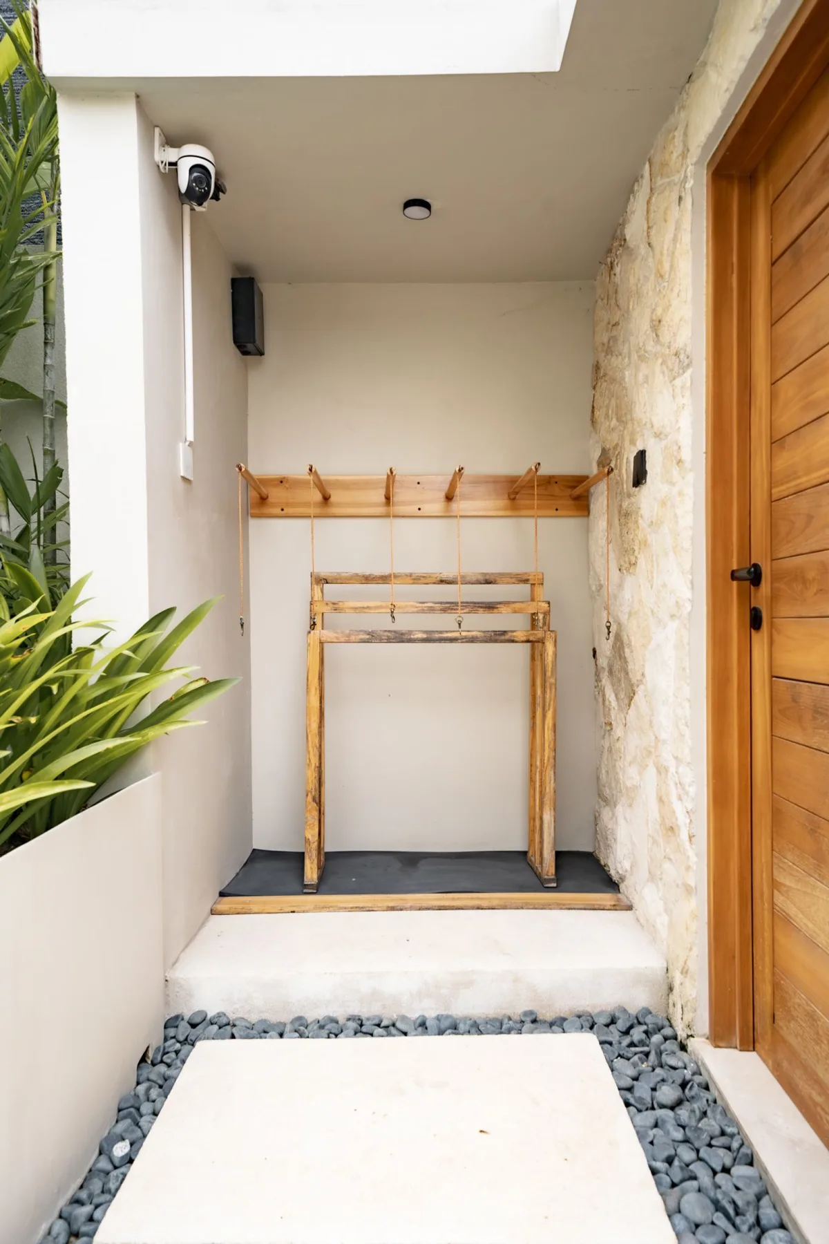 A narrow covered entryway has a wooden door, stone accent wall, and a simple wooden luggage rack.