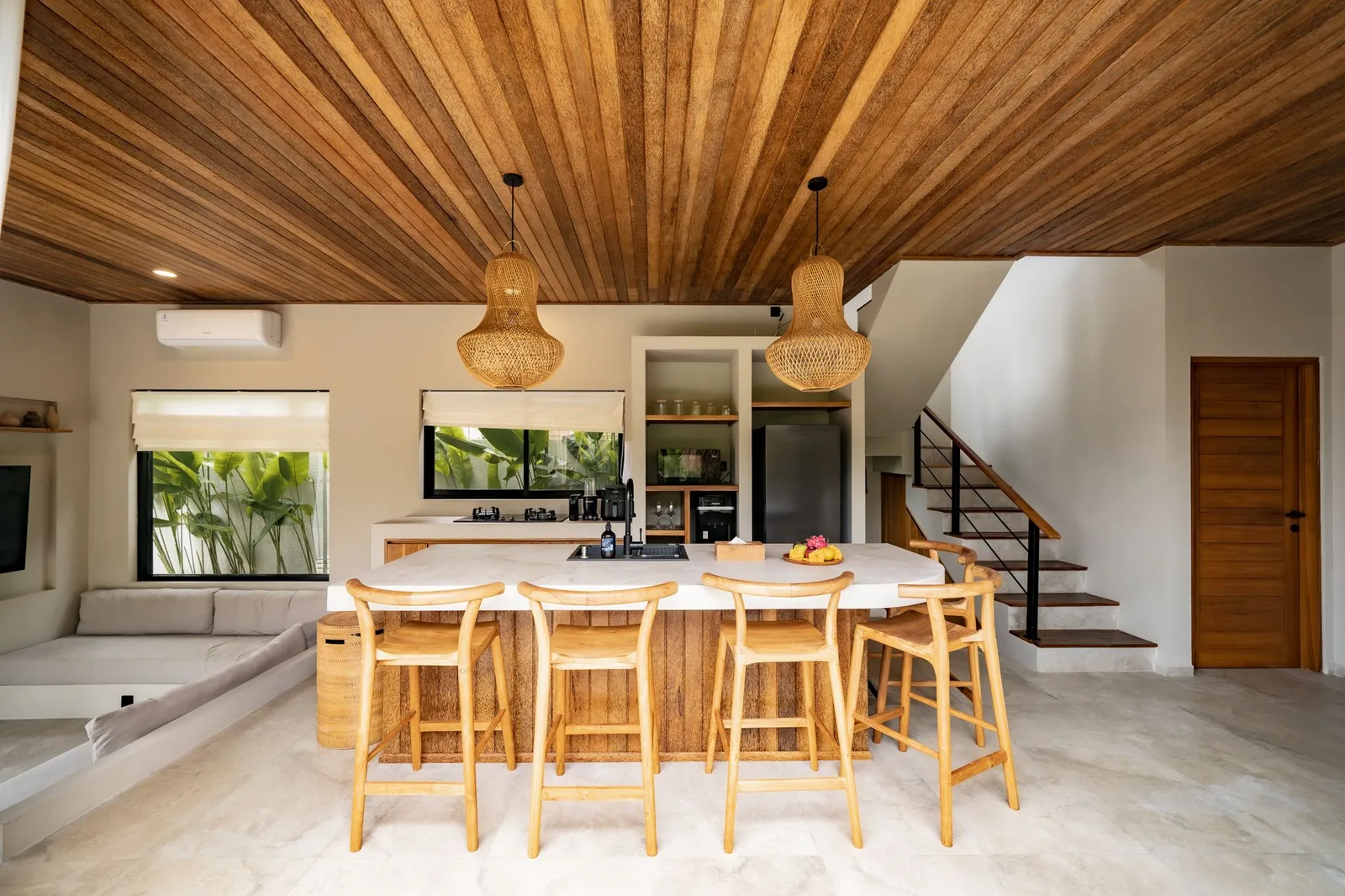 A bright open-plan dining area features a long wooden table, woven pendant lights, and stairs beside a wooden door.
