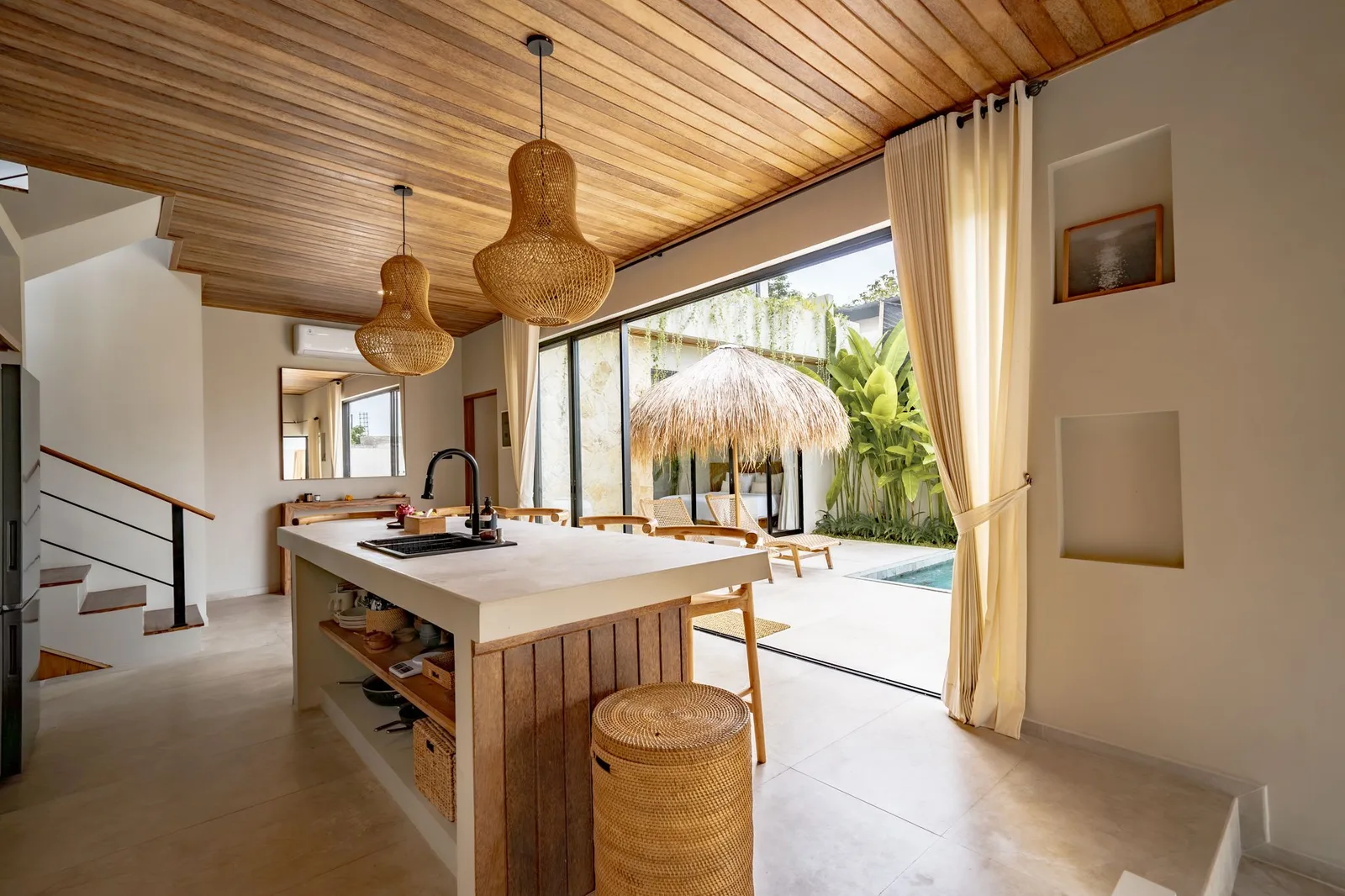 An open kitchen with a long island, woven pendant lights, and sliding doors to a tropical patio.