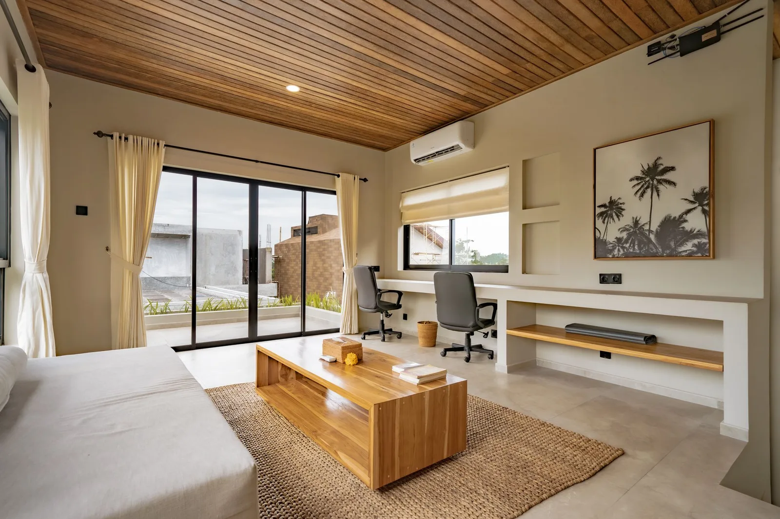A bright living room has a sofa, wooden coffee table, desk, and sliding glass doors beneath a timber ceiling.