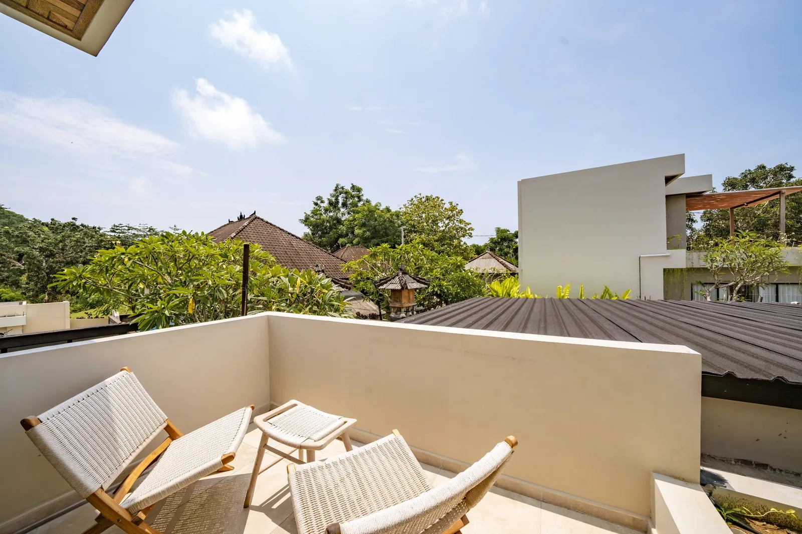 A sunny rooftop terrace has woven lounge chairs, a small table, and views of nearby rooftops and trees.
