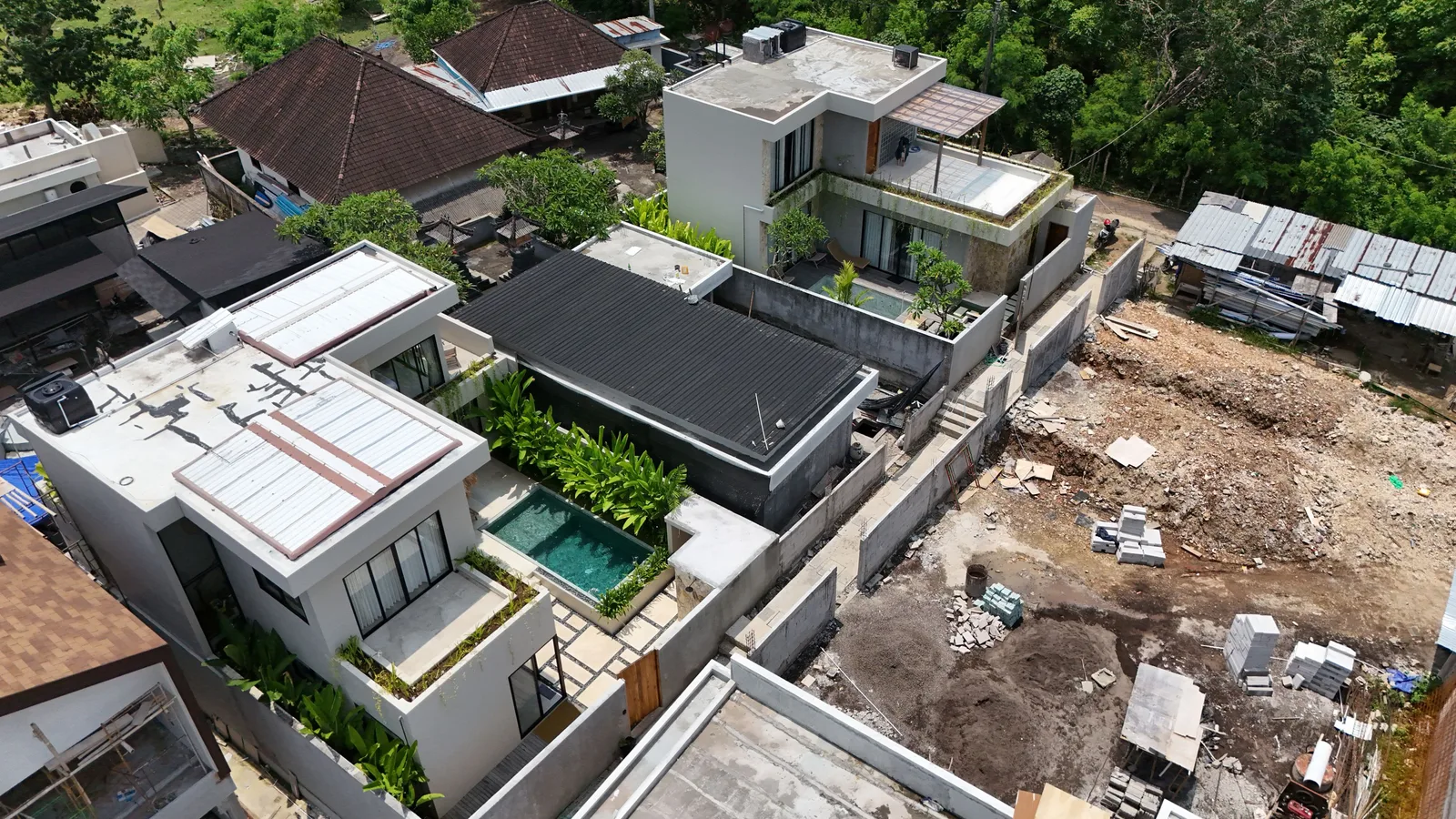 An aerial view shows modern flat-roofed homes beside a cleared construction lot surrounded by trees.