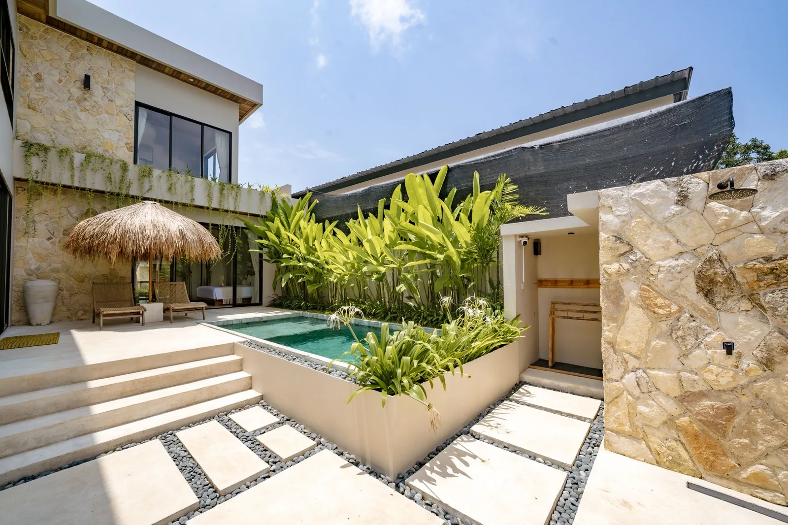 Modern stone house with a small courtyard pool, tropical plants, and a thatched patio umbrella.